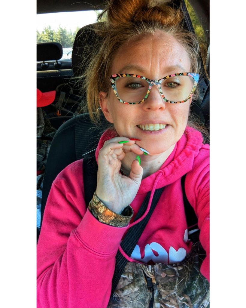A smiling woman with colorful glasses and a bright pink hoodie, sitting in a car. She is playfully touching her face, showcasing her stylish nails and casual attire.