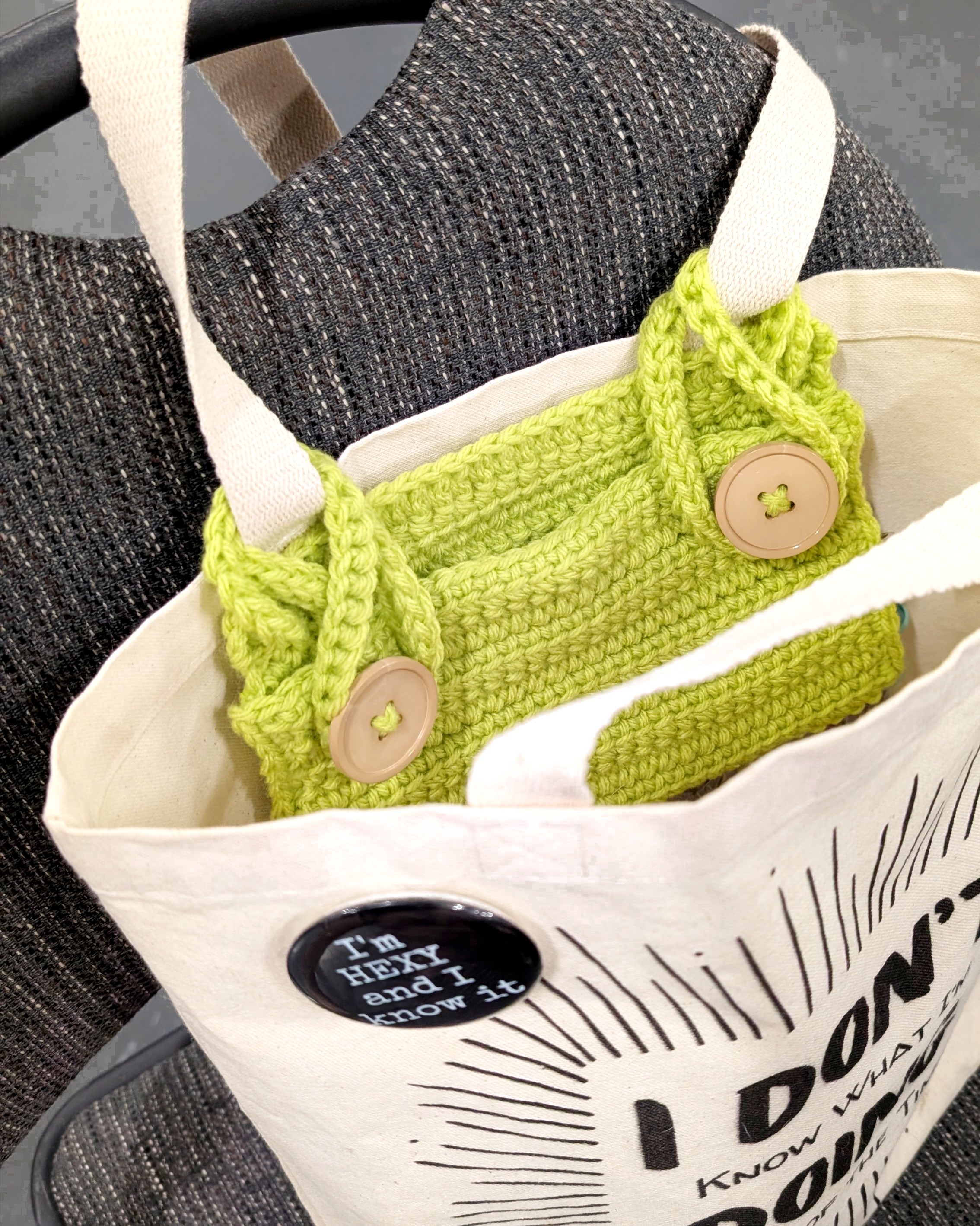 Close-up of a canvas tote bag displaying a humorous pin and a crocheted green item inside.