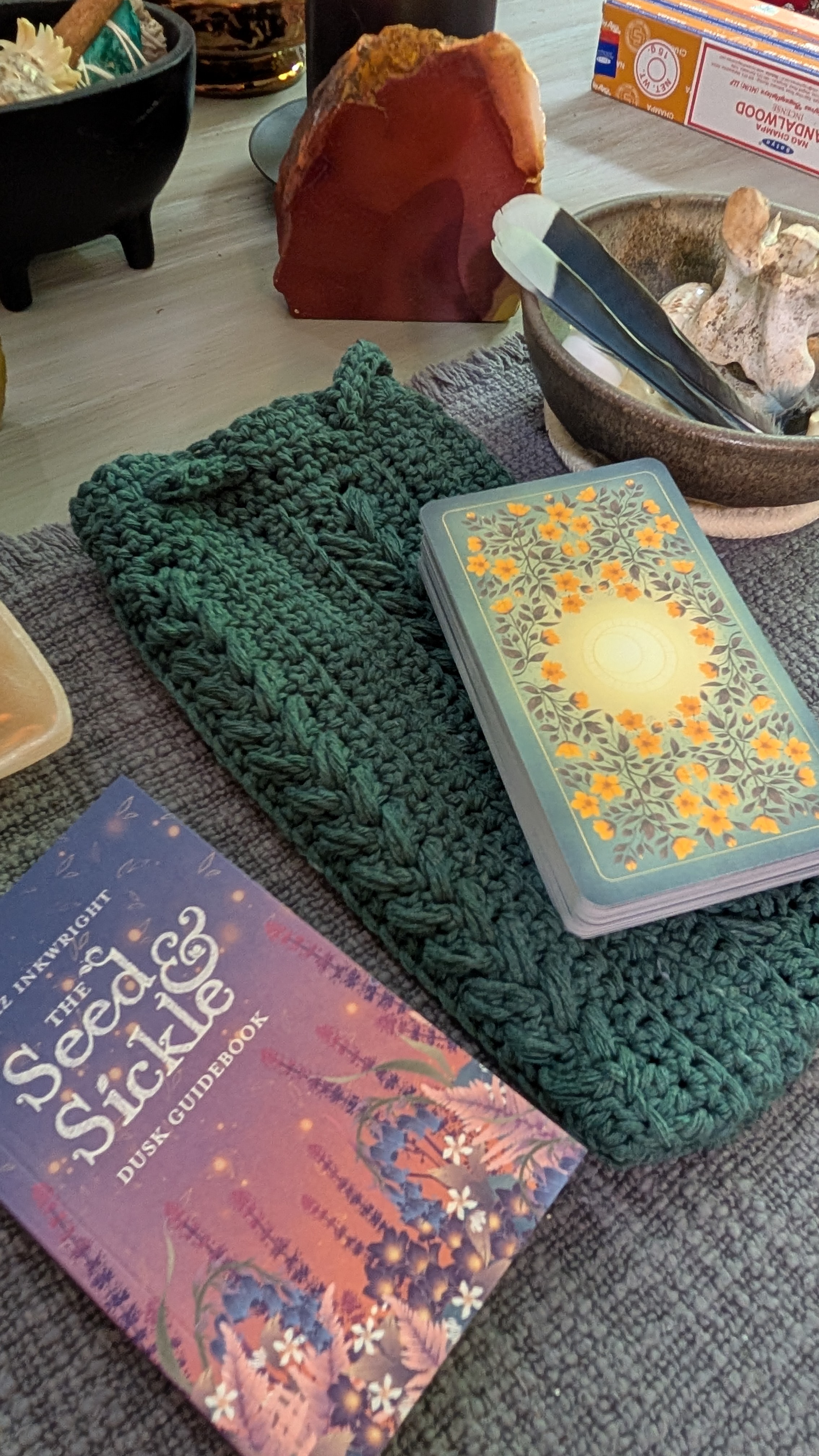 A crochet item in green, a book titled 'The Seed & Sickle Dusk Guidebook', and a deck of cards with a floral design, arranged on a textured surface.